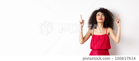 Dreamy young woman in trendy red dress, looking up and pointing at logo, smiling happy, standing over white background Dreamy young woman in trendy red dress, looking up and pointing at logo, smiling happy, standing over white background 100617614