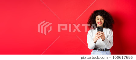Smiling modern girl taking photos on smartphone, looking at screen and recording video, standing against red background Smiling modern girl taking photos on smartphone, looking at screen and recording video, standing against red background 100617696