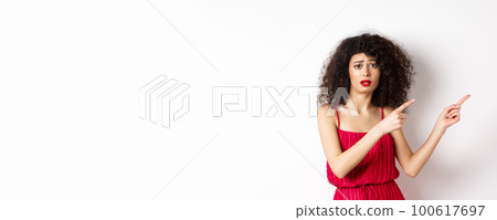 Confused and sad woman with curly hair, wearing evening dress and makeup, pointing fingers right and frowning upset, standing on white background Confused and sad woman with curly hair, wearing evening dress and makeup, pointing fingers right and frowning upset, standing on white background 100617697