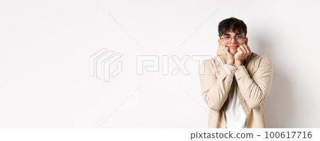 Portrait of handsome caucasian man watching something lovely and cute, squeeze cheeks and lean on hands, looking with admiration and interest at camera, standing on white background 100617716