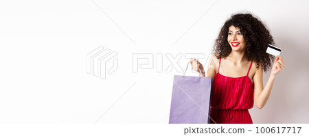 Beautiful woman with curly hair, red dress, showing shopping bag and plastic credit card, white background Beautiful woman with curly hair, red dress, showing shopping bag and plastic credit card, white background 100617717