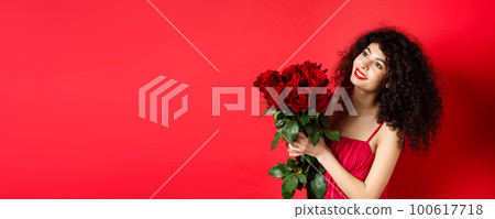 Happy beautiful woman in dress, holding flowers and smiling romantic, looking aside at logo, standing against red background Happy beautiful woman in dress, holding flowers and smiling romantic, looking aside at logo, standing against red background 100617718