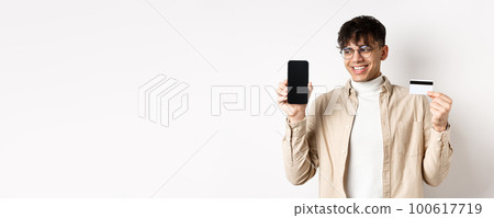 Online shopping. Handsome guy in glasses showing empty cellphone screen and credit card, smiling and looking at display pleased, standing on white background Online shopping. Handsome guy in glasses showing empty cellphone screen and credit card, smiling and looking at display pleased, standing on white background 100617719