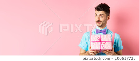 Holidays and celebration concept. Funny guy with moustache showing cute gift box wrapped in festive paper, looking at camera, presenting you, standing on pink background 100617723