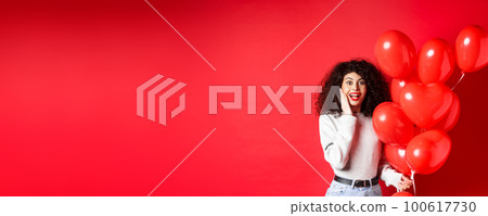 Valentines day. Surprised curly-haired girl holding heart balloons and looking amazed at camera, receive surprise gift on date from lover, standing on red background 100617730