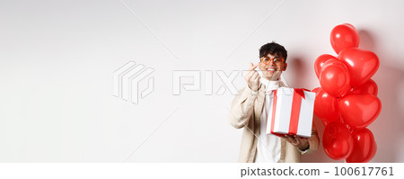 Valentines day and romance concept. Handsome young man showing surprise gift for lover and finger heart, smiling, standing near festive red balloons, white background Valentines day and romance concept. Handsome young man showing surprise gift for lover and finger heart, smiling, standing near festive red balloons, white background 100617761