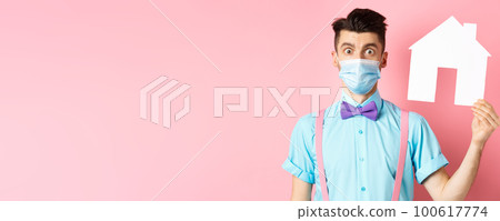 Covid, pandemic and real estate concept. Confused guy in medical mask searching property, showing house cutout and stare at camera worried, standing over pink background Covid, pandemic and real estate concept. Confused guy in medical mask searching property, showing house cutout and stare at camera worried, standing over pink background 100617774