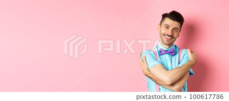 Cheerful young man smiling and hugging himself, wrap hands around own body and looking happy, standing over pink background 100617786