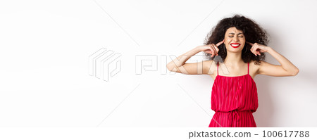 Annoyed woman with curly hair and red dress, shut eyes and ears, block annoying sound, clench teeth bothered, complain on loud music or neighbours, standing on white background 100617788