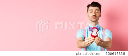 Valentines day. Silly boyfriend in bow-tie pucker lips, close eyes and waiting for kiss after making proposal, showing golden engagement ring in small red box, standing on pink background 100617836