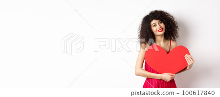 Romantic tender woman with curly hair, hugging big red heart and smiling, look with love, standing against white background 100617840