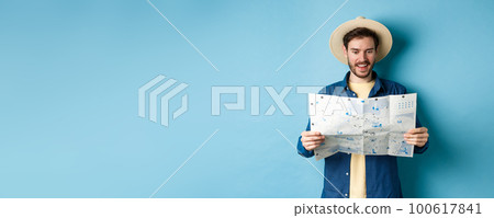 Cheerful tourist in summer hat planning travel route on vacation, looking at sighseeing map and smiling excited, standing on blue background 100617841
