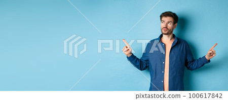 Indecisive man in casual clothes making choice, pointing fingers sideways and looking pensive, standing on blue background 100617842