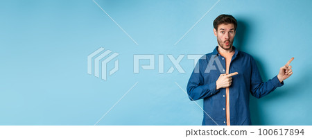 Surprised young man checking out promo, pointing fingers right at logo and saying wow, standing on blue background Surprised young man checking out promo, pointing fingers right at logo and saying wow, standing on blue background 100617894