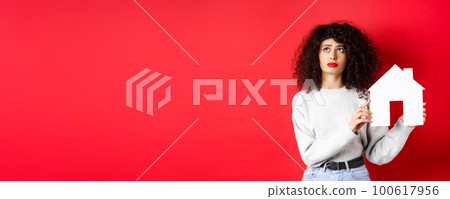 Real estate. Sad woman thinking of buying own house, showing paper home cutout and looking up with uspet face, standing on red background Real estate. Sad woman thinking of buying own house, showing paper home cutout and looking up with uspet face, standing on red background 100617956