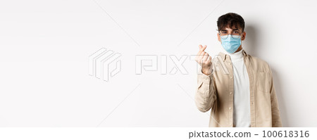 Covid-19, health and real people concept. Cute young man in medical mask showing finger heart and look at camera, standing on white background Covid-19, health and real people concept. Cute young man in medical mask showing finger heart and look at camera, standing on white background 100618316