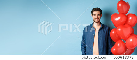 Cute boyfriend looking happy and smiling, standing near Valentines day heart balloons on blue background Cute boyfriend looking happy and smiling, standing near Valentines day heart balloons on blue background 100618372