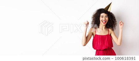 Birthday girl in party hat having fun, dancing in red dress and chanting, standing against white background Birthday girl in party hat having fun, dancing in red dress and chanting, standing against white background 100618391