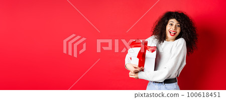 Valentines day. Happy girlfriend holding gift box from secret admirer, looking romantic at lover, receive present, standing on red background 100618451