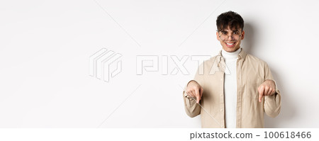 Stylish hipster male model in glasses pointing fingers down, smiling pleased at camera, recommending product, standing on white background Stylish hipster male model in glasses pointing fingers down, smiling pleased at camera, recommending product, standing on white background 100618466