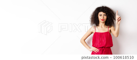 Smiling caucasian woman with curly hair, wearing red dress, showing rule number one gesture, raising finger and looking confident at camera, white background Smiling caucasian woman with curly hair, wearing red dress, showing rule number one gesture, raising finger and looking confident at camera, white background 100618510