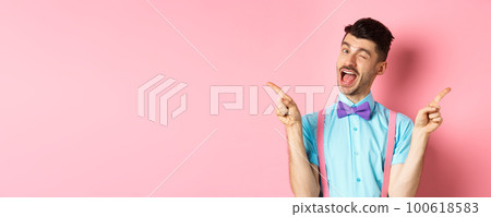 Cheerful young man smiling and winking, showing two sides, pointing fingers sideways and recommending promo offers on pink background 100618583