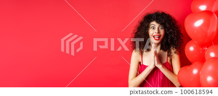 Valentines day. Excited caucasian woman in dress jumping from amazement, looking at something taunting, want to get product, standing near hearts balloons, white background 100618594