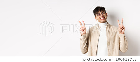 Handsome hipster guy in glasses, smiling with white teeth, showing peace or victory gesture, standing upbeat on white background 100618713
