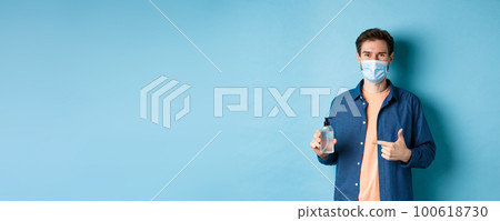 Coronavirus, quarantine and social distancing concept. Happy young guy in medical mask recommending bottle of hand sanitizer, pointing at antiseptic and smiling, blue background Coronavirus, quarantine and social distancing concept. Happy young guy in medical mask recommending bottle of hand sanitizer, pointing at antiseptic and smiling, blue background 100618730