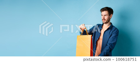 Handsome young man, shopping salesman stretching out orange shopping bag to client, looking aside, standing on blue background 100618751