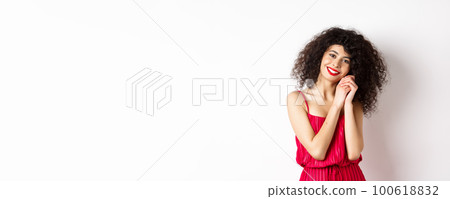 Lovely young woman with curly hairstyle, wearing red dress, looking at silly and tender thing, admire something, standing over white background 100618832