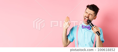 Party and festive events concept. Funny guy performs a song, singing karaoke in microphone with carefree face, standing in bow-tie and suspenders on pink background Party and festive events concept. Funny guy performs a song, singing karaoke in microphone with carefree face, standing in bow-tie and suspenders on pink background 100618852