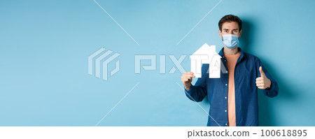 Real estate and covid concept. Young man in face mask showing paper house cutout and thumb-up, recommending agency, standing on blue background Real estate and covid concept. Young man in face mask showing paper house cutout and thumb-up, recommending agency, standing on blue background 100618895