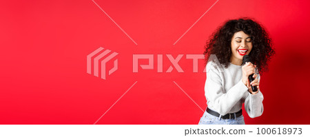 Hobbies and leisure concept. Beautiful woman singing in microphone and smiling, having fun at karaoke, standing on red background 100618973
