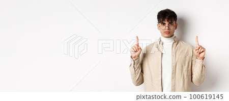 Handsome hipster guy in glasses pointing fingers up at empty space, looking serious at camera, standing on white background Handsome hipster guy in glasses pointing fingers up at empty space, looking serious at camera, standing on white background 100619145