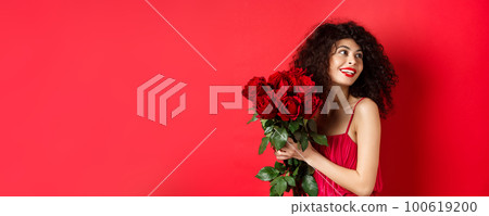Tender young woman in elegant red dress, holding romantic bouquet of red roses and looking right, smiling dreamy, thinking about lover on Valentines day, studio background 100619200
