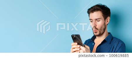 Image of impessed young man looking at smartphone and saying wow, checking out online offer, standing on blue background Image of impessed young man looking at smartphone and saying wow, checking out online offer, standing on blue background 100619885