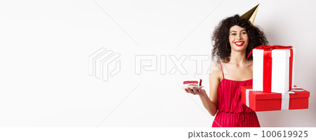 Happy birthday girl in red dress, celebrating and holding gifts with bday cake, standing on white background 100619925