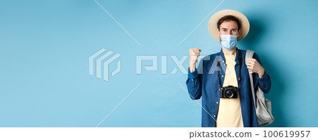 Covid-19 and summer holidays concept. Happy young man travelling abroad in medical mask, saying yes and raising fist up joyfully, cheerful about vacation, blue background Covid-19 and summer holidays concept. Happy young man travelling abroad in medical mask, saying yes and raising fist up joyfully, cheerful about vacation, blue background 100619957