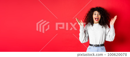 Image of happy and surprised curly woman in makeup and sweatshirt, raising hands up and rejoicing from good news, standing on red background Image of happy and surprised curly woman in makeup and sweatshirt, raising hands up and rejoicing from good news, standing on red background 100620078
