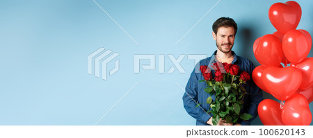 Romantic boyfriend winking and smiling, holding bouquet of flowers on Valentines day, standing near heart balloons for lover, blue background 100620143