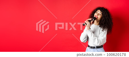 Leisure and hobbies concept. Stylish young woman singing karaoke, looking aside and holding microphone, performing song, standing on red background Leisure and hobbies concept. Stylish young woman singing karaoke, looking aside and holding microphone, performing song, standing on red background 100620178