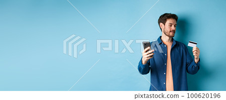 E-commerce concept. Man paying for online payment, reading credit card number and holding mobile phone, standing on blue background E-commerce concept. Man paying for online payment, reading credit card number and holding mobile phone, standing on blue background 100620196