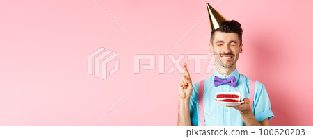 Holidays and celebration concept. Hopeful birthday guy in party hat making wish, holding cake with candle and cross fingers for dream come true, pink background Holidays and celebration concept. Hopeful birthday guy in party hat making wish, holding cake with candle and cross fingers for dream come true, pink background 100620203