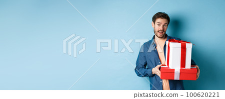 Handsome bearded guy holding Valentines day gifts for lover, standing with presents in boxes and looking at camera, blue background Handsome bearded guy holding Valentines day gifts for lover, standing with presents in boxes and looking at camera, blue background 100620221