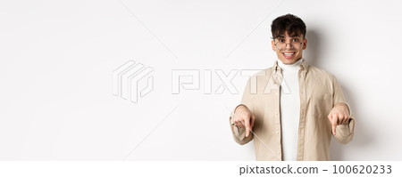Portrait of happy young guy in glasses showing cool promo, pointing fingers down and smiling excited, standing on white background Portrait of happy young guy in glasses showing cool promo, pointing fingers down and smiling excited, standing on white background 100620233