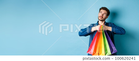 Dreamy young man holding colorful shopping bags and imaging something, looking at upper left corner empty space, blue background Dreamy young man holding colorful shopping bags and imaging something, looking at upper left corner empty space, blue background 100620241