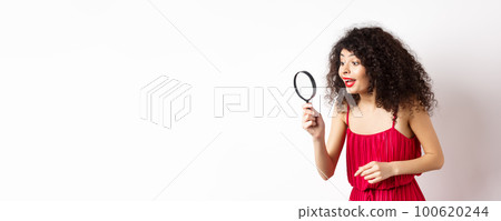 Beautiful woman in red dress and makeup looking at something with magnifying glass, checking out interesting promo, standing over white background Beautiful woman in red dress and makeup looking at something with magnifying glass, checking out interesting promo, standing over white background 100620244