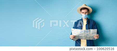 Covid-19, pandemic and travel concept. Young man in medical mask reading tourist map, searching for route to sightseeings, standing on blue background 100620516