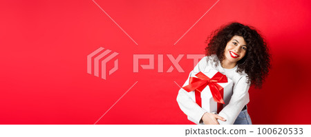 Celebration and holidays concept. Happy woman holding birthday gift and smiling at camera, standing in casual clothes, red background 100620533